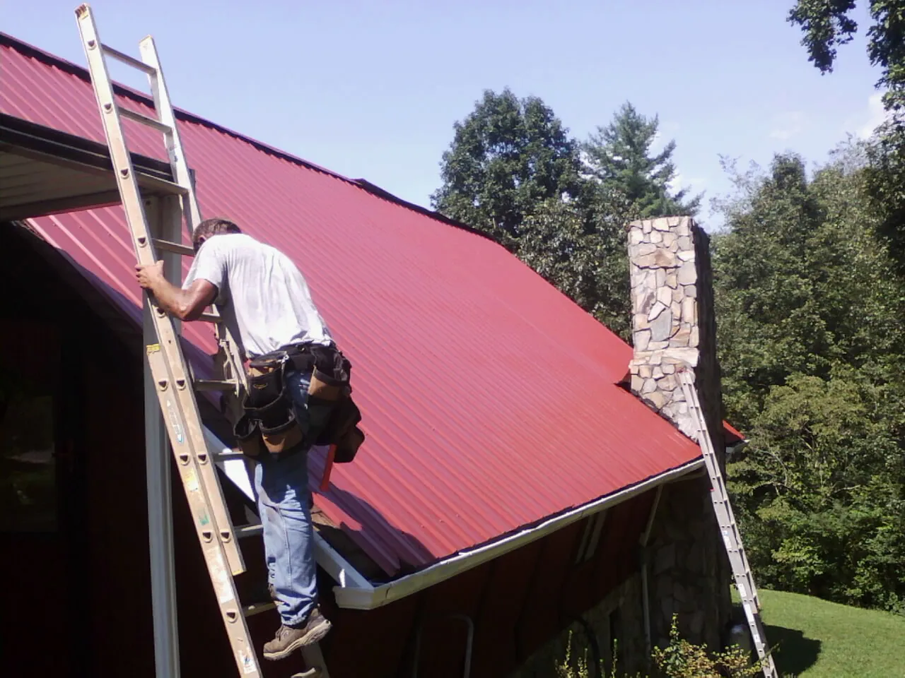 Roofer on a ladder beside a red metal roof and stone chimney — Soffit & Fascia Repair by Roofing Brothers in Lakeside Marblehead